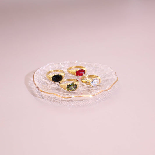 Four gold rings with different colored stones are arranged on a small clear glass plate against a soft pink background.