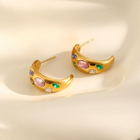 A pair of gold crescent-shaped hoop earrings decorated with colorful gemstones, including pink, green, purple, and clear stones, displayed on a warm cream-colored surface.