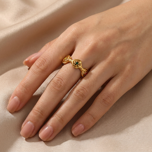 A gold ring with gemstones and decorative side details, shown worn on a hand resting on a beige satin fabric background.