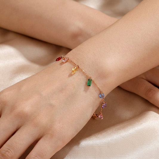 A delicate gold chain bracelet with small dangling colorful gemstones, shown worn on a person's wrist against a soft beige satin background.