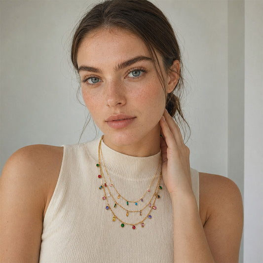 A woman wearing a delicate gold necklaces with small colorful gemstone charms, paired with a cream ribbed mock neck top, set against a soft neutral background.