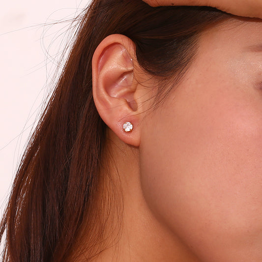 A close-up of a woman's ear wearing a small round stud earring with a clear sparkling gemstone on a light pink background.
