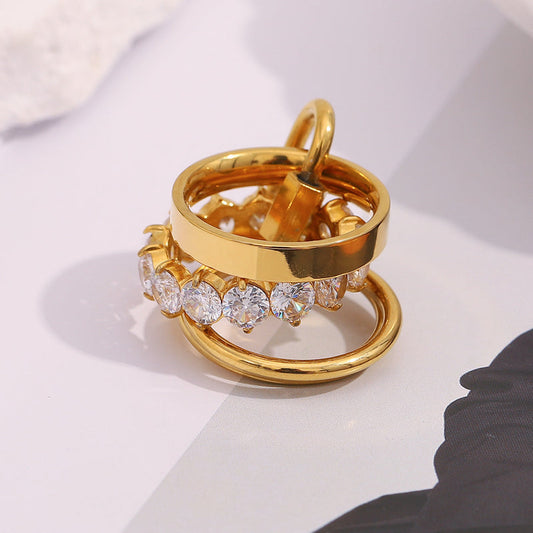 A stack of gold rings, including one with a row of clear round-cut stones, photographed on a light gray surface with soft shadows.