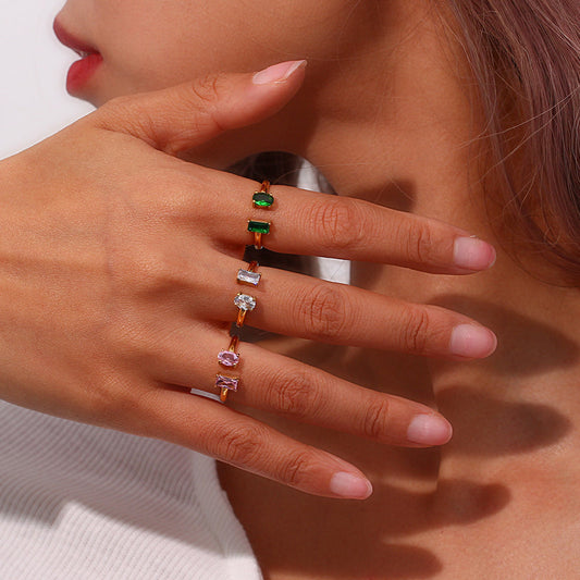 A close-up of a person's hand wearing a stack of slim gold rings, each set with a different colored gemstone (green, clear, pink).