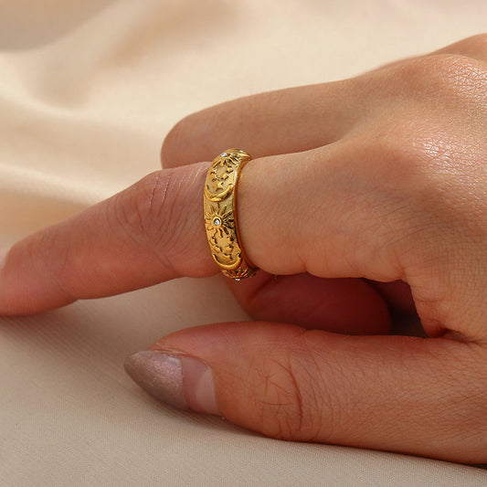 A close-up of a hand wearing a textured gold ring with a small clear stones, resting against a soft beige fabric background.