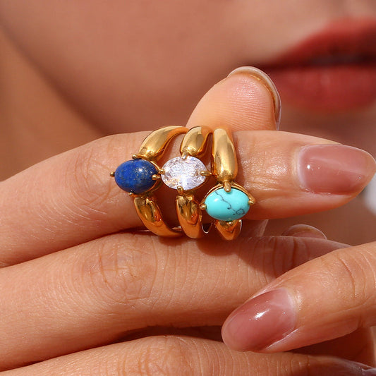 A close-up of gold rings with three stacked bands, featuring a blue stone, a clear oval stone, and a turquoise stone, worn on a person's finger.