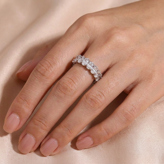 A close-up of a hand wearing a silver-toned eternity ring set with clear round-cut gemstones, resting on a soft beige satin fabric background.