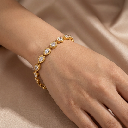 A gold bracelet with teardrop-shaped clear gemstones set in gold, worn on a person's wrist against a soft beige satin background.