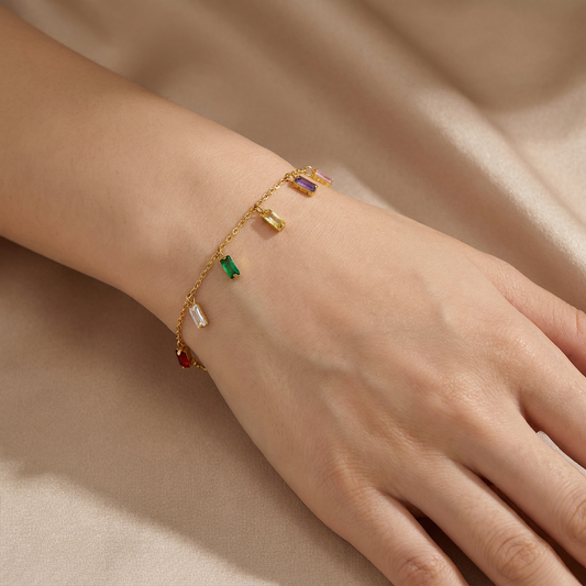A delicate gold chain bracelet with small colorful rectangular charms, worn on a person's wrist against a soft beige satin background.