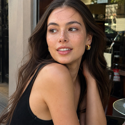 A woman with long dark brown hair wearing a black ribbed tank top and gold hoop earrings, looking to the side in an outdoor cafe setting.
