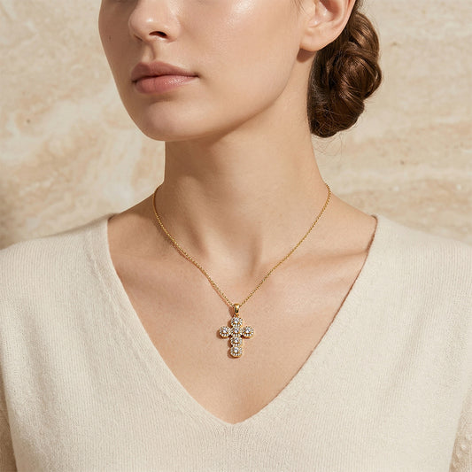 A close-up of a woman wearing a delicate gold cross pendant necklace adorned with small clear crystals, styled with a cream-colored V-neck sweater against a textured beige background.