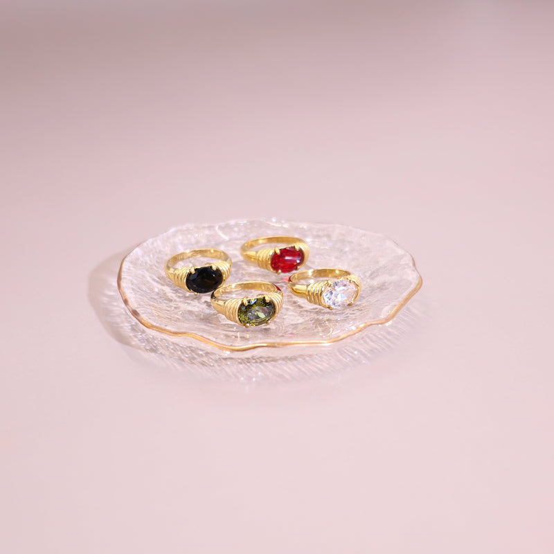 Four gold rings with different colored stones are arranged on a small clear glass plate against a soft pink background.