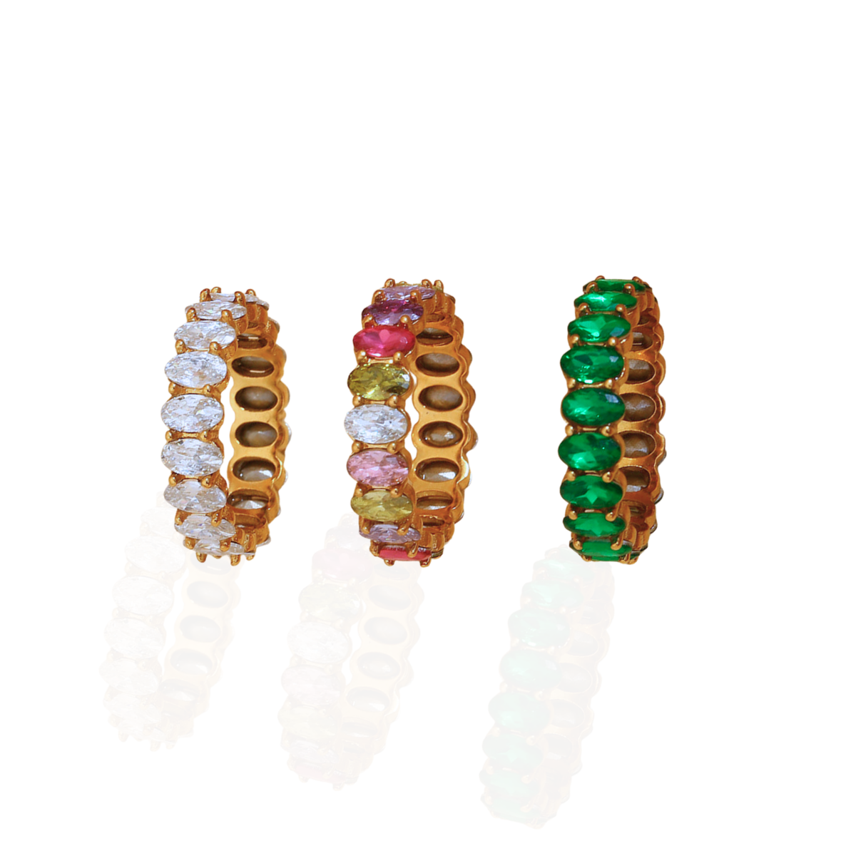 Three gold-plated stretch rings with oval-cut gemstones are displayed side-by-side on a white reflective surface. The left ring features clear stones, the middle ring features pink, yellow, and brown stones, and the right ring features green stones.