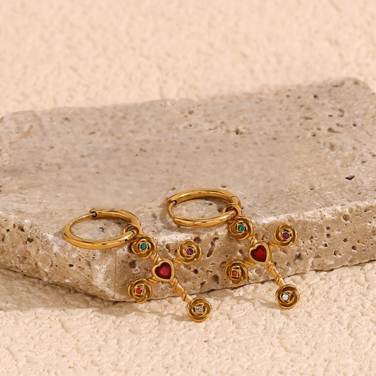 A pair of gold hoop earrings with dangling chain-like links and small colorful gemstone accents, displayed on a textured beige stone slab against a matching beige background.
