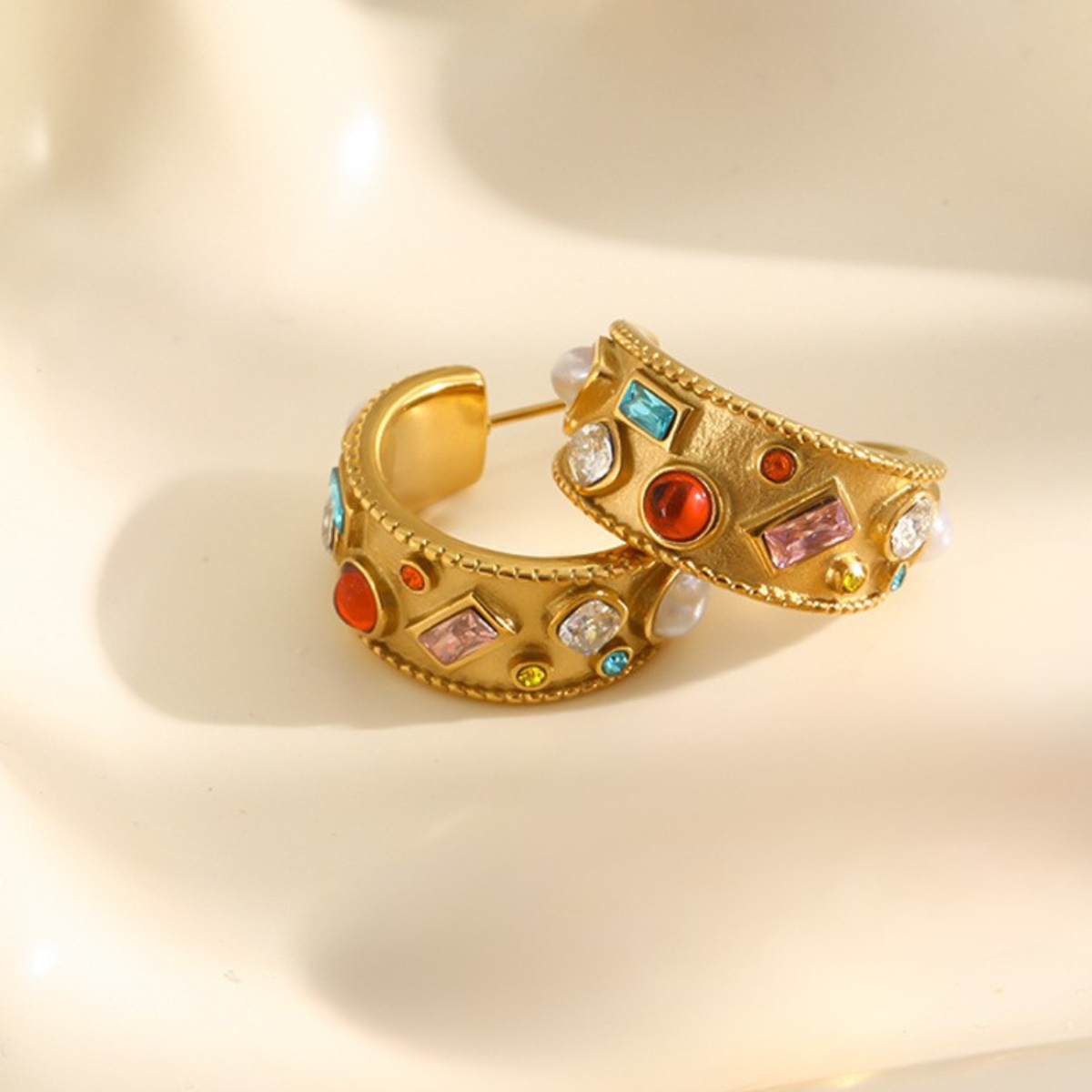 A pair of gold hoop earrings decorated with colorful faux gemstones and white faux pearls, resting on a warm beige surface.