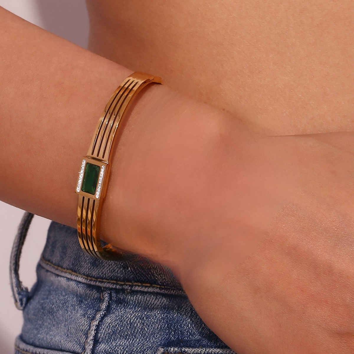 A gold-toned multi-band bracelet with a central green rectangular stone, worn on a person's wrist above blue denim jeans.