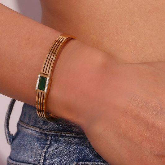 A gold-toned multi-band bracelet with a central green rectangular stone, worn on a person's wrist above blue denim jeans.