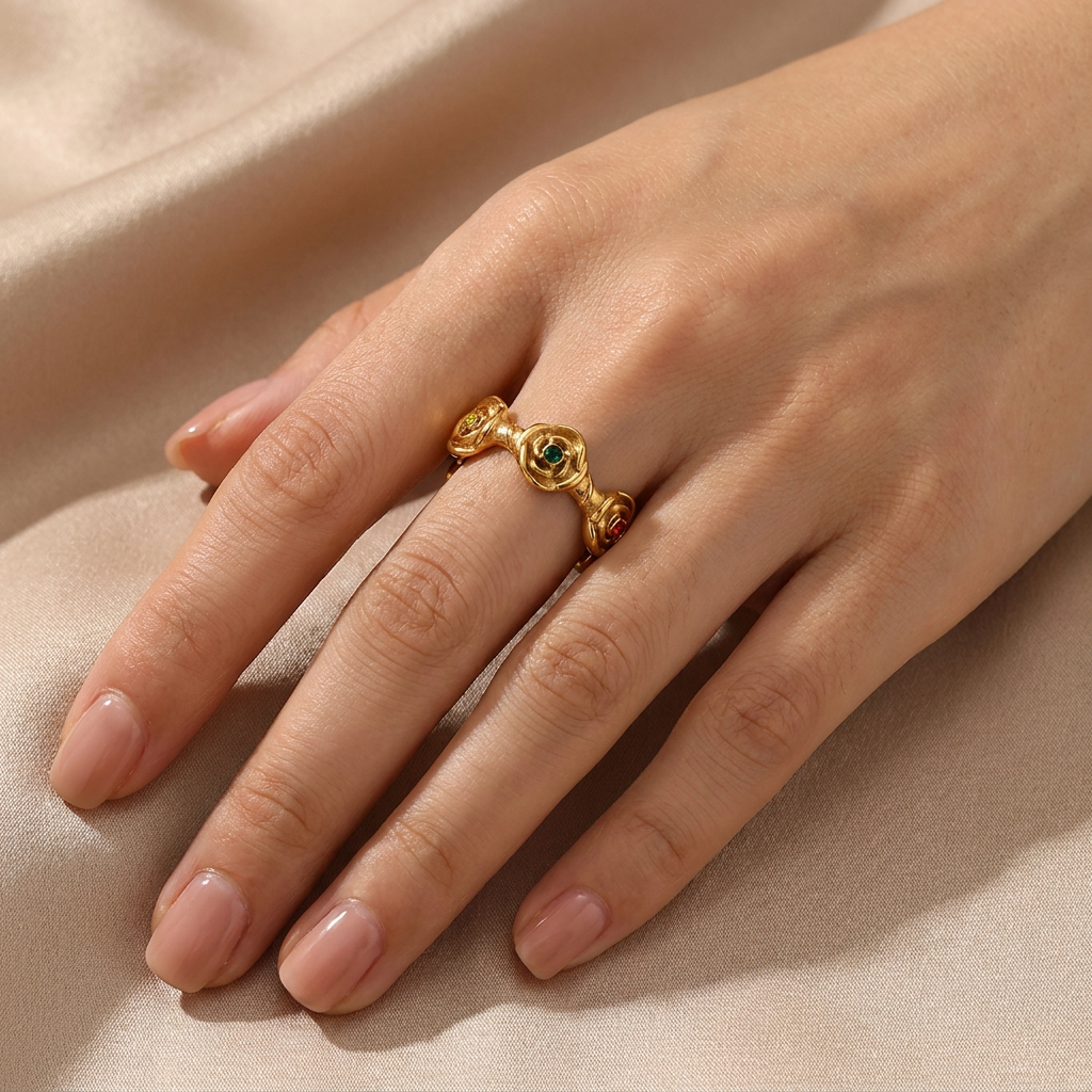 A gold ring with gemstones and decorative side details, shown worn on a hand resting on a beige satin fabric background.