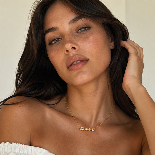 A close-up lifestyle shot of a woman with long dark brown hair wearing a delicate gold necklace with three small, round, pearl-like beads on a bar. She is wearing a white off-the-shoulder top against a soft, light beige background.
