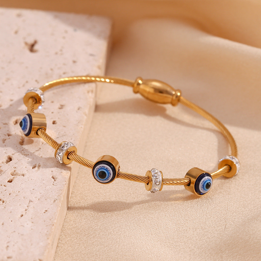Gold-plated bangle bracelet with evil eye charms and silver crystal accents, displayed on a beige fabric and stone surface.