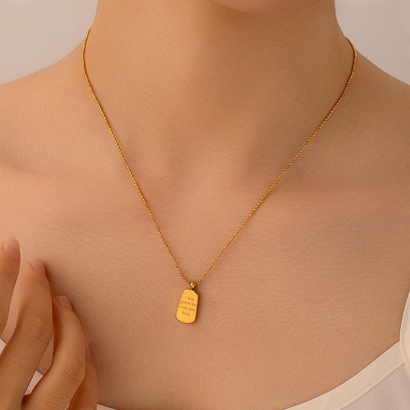 A close-up of a model's neck and chest wearing a delicate gold chain necklace with a small rectangular gold pendant engraved with text, set against a soft beige background.
