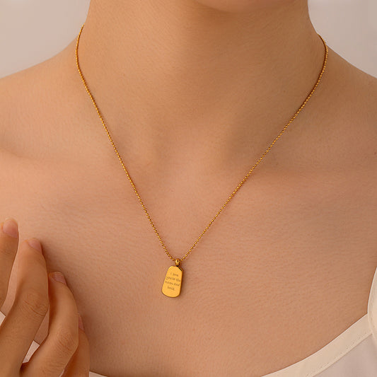 A close-up of a model's neck and chest wearing a delicate gold chain necklace with a small rectangular gold pendant engraved with text, set against a soft beige background.