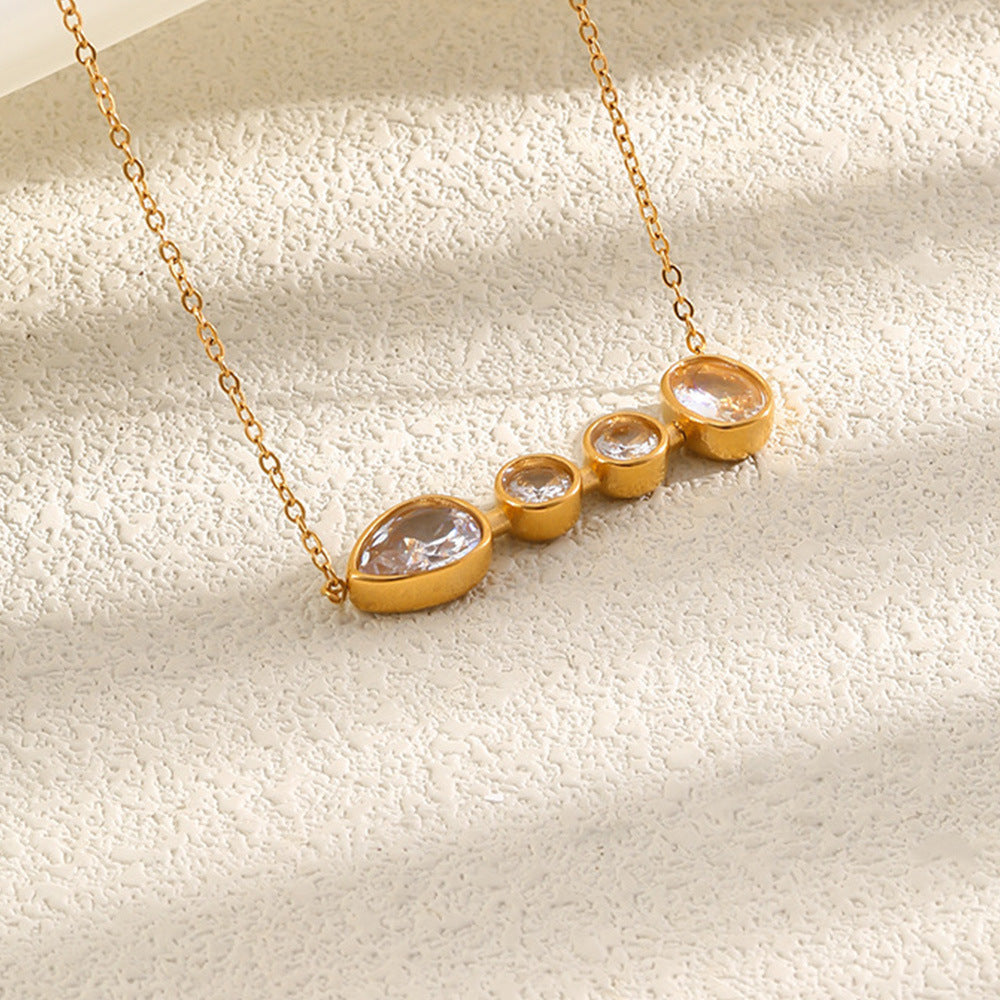 Gold necklace with a horizontal bar featuring three bezel-set clear teardrop-shaped gemstones of decreasing size, displayed on a textured cream surface with soft diagonal shadows.