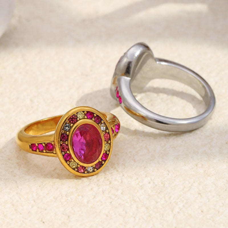 Two rings on a textured beige surface: one gold ring with an oval pink gemstone surrounded by small pink and clear stones, and one silver ring shown from the side.