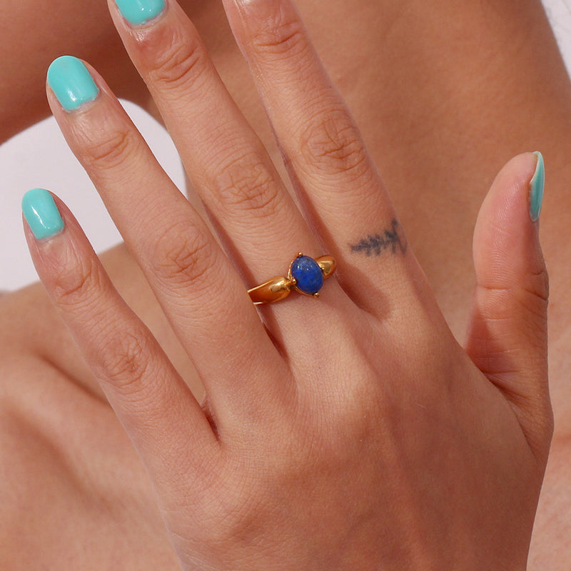 A gold ring with a round blue gemstone worn on a person's finger, shown in a close-up lifestyle shot with turquoise nail polish.