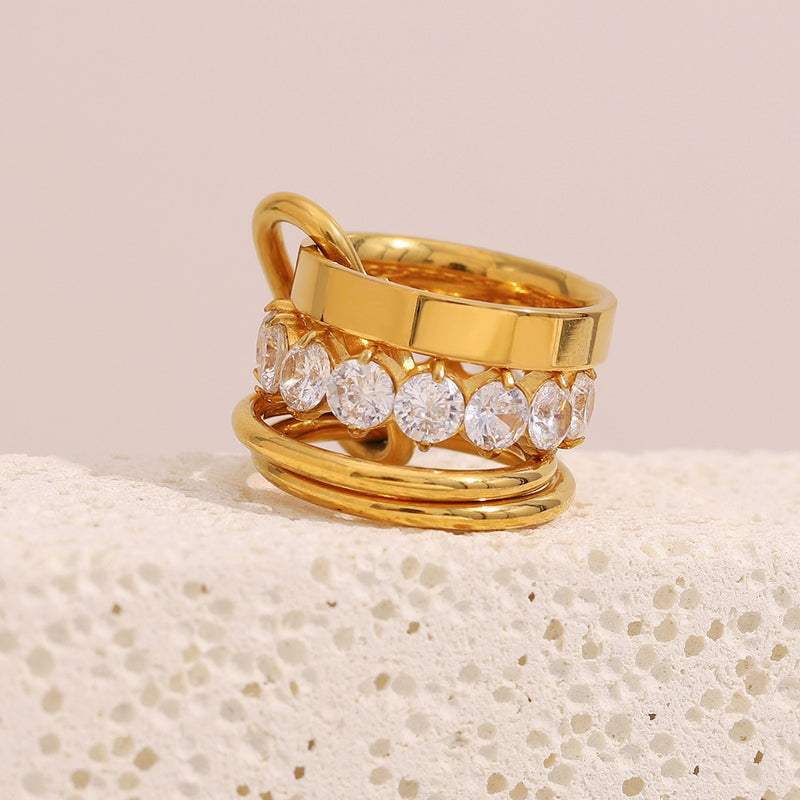 A gold-plated multi-band ring featuring a row of clear round-cut gemstones on one band, displayed on a textured white surface against a soft beige background.
