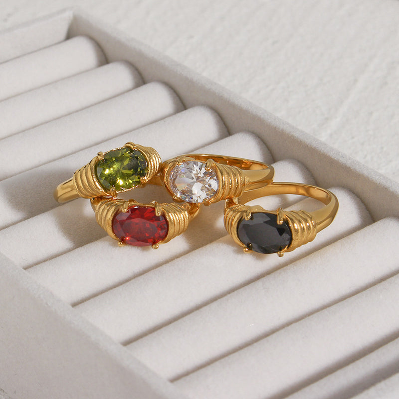 Four gold rings with oval gemstones in green, red, clear, and black, displayed on a white ring box.