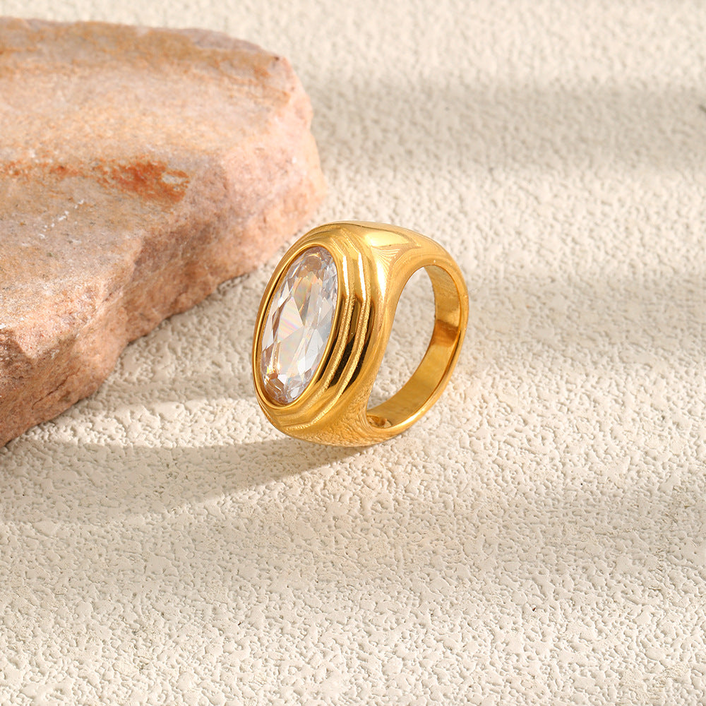 A gold signet-style ring with a large oval clear gemstone, resting on a textured beige surface next to a pink stone.