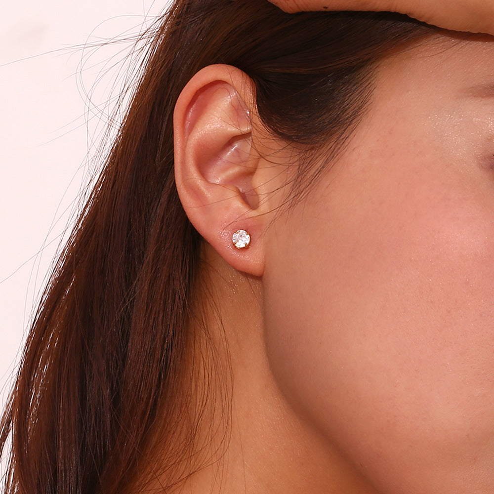 A close-up of a woman's ear wearing a small round stud earring with a clear sparkling gemstone on a light pink background.