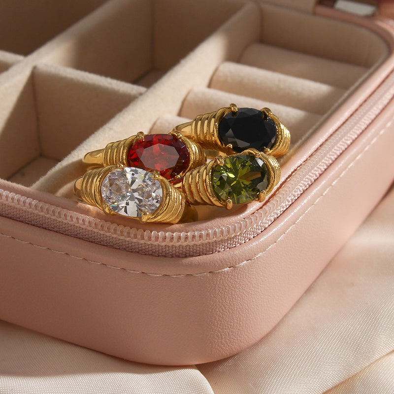 Three gold-toned ring-shaped earrings with round gemstones in red, black, clear, and green settings, displayed inside a light pink jewelry organizer pouch.