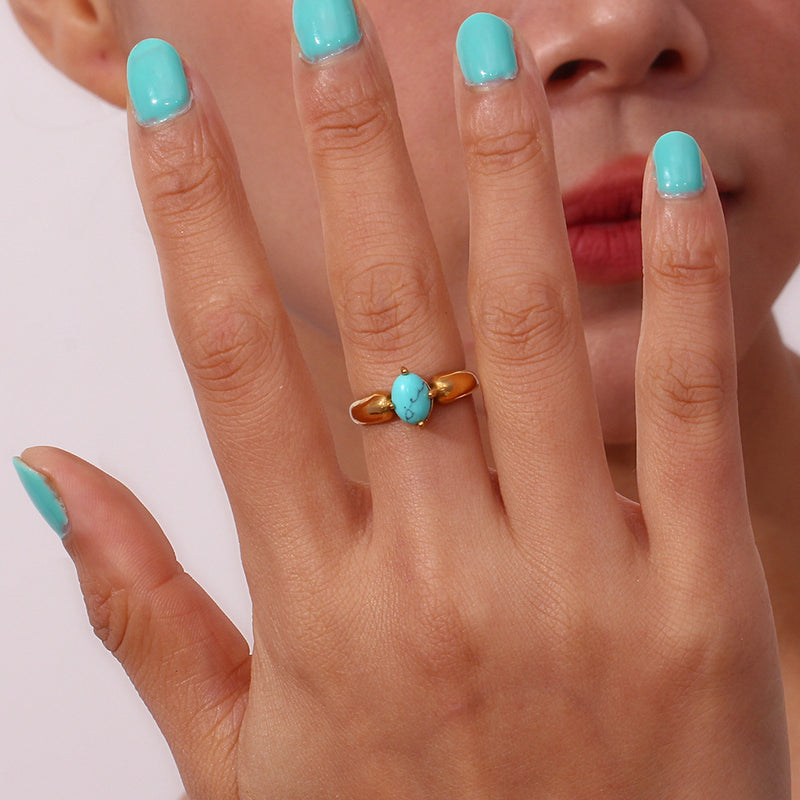 A close-up of a hand with light blue painted nails wearing a gold ring with a round turquoise cabochon stone, with part of a person's face visible in the background.