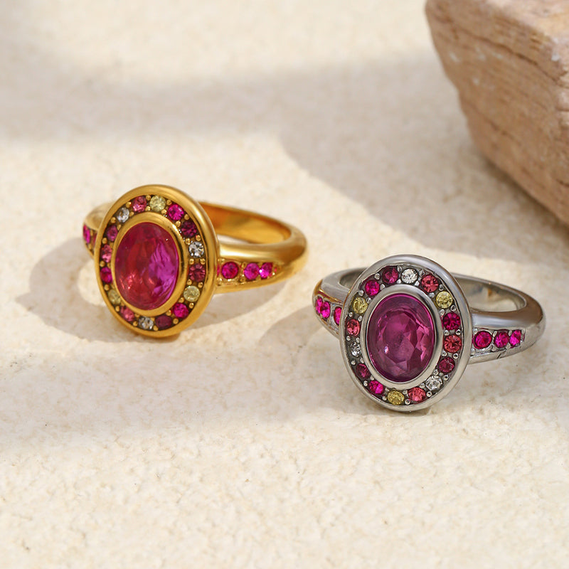 Two oval gemstone rings displayed on a light textured surface. One ring is gold-toned with a magenta center stone and pink and yellow accent stones.