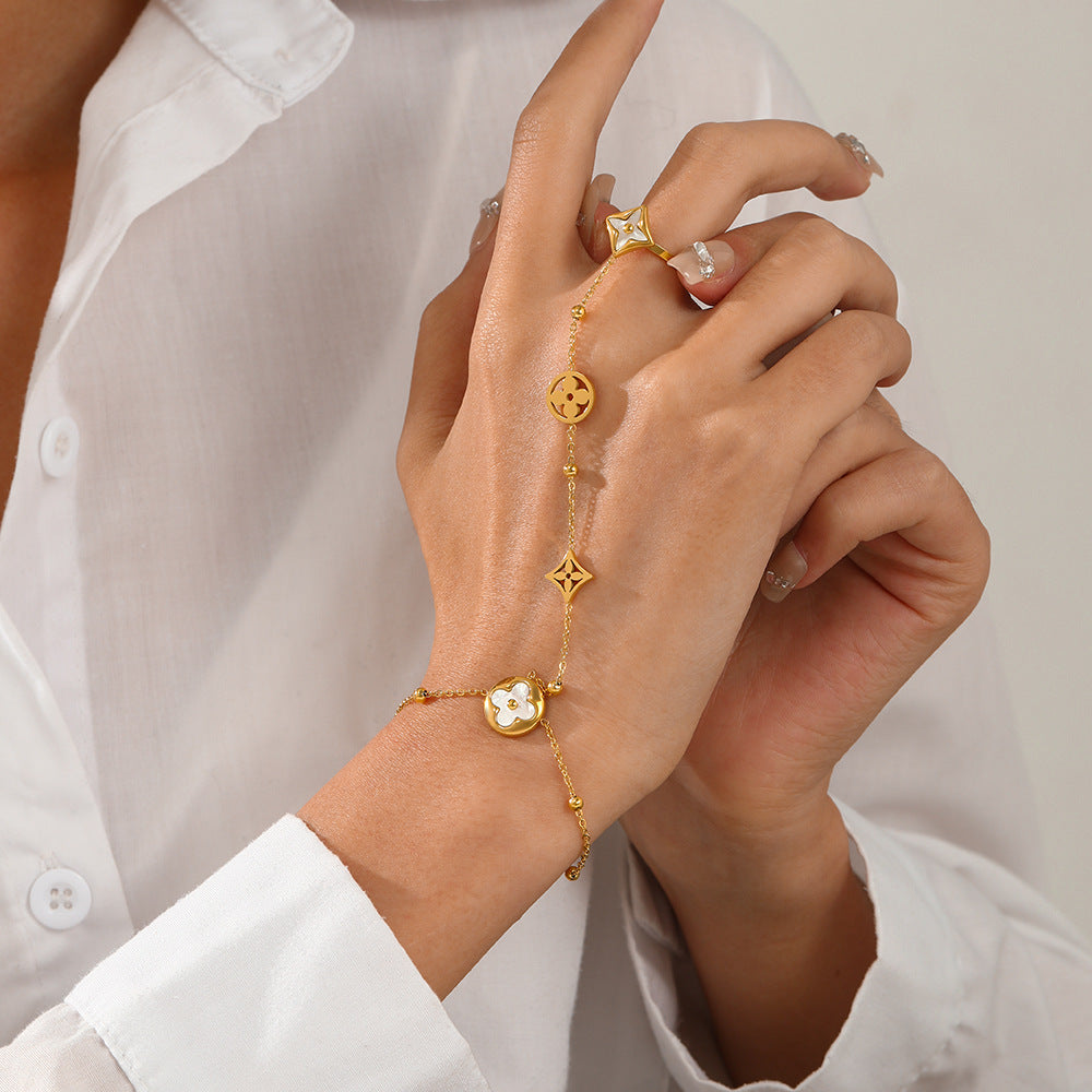 A close-up of a model's hands wearing a delicate gold chain bracelet with a small oval charm and a matching gold ring, styled with a white button-down shirt.