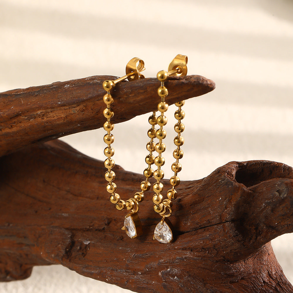 A pair of gold beaded drop earrings with small clear teardrop crystals, displayed on a dark brown wooden branch against a soft off-white background.