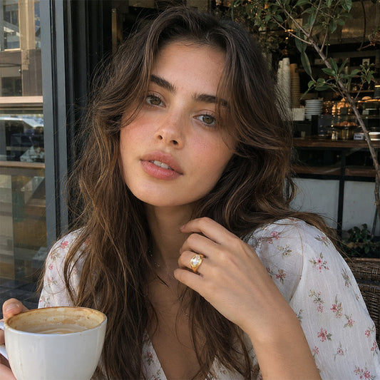 A woman with long wavy brown hair is shown in a cafe setting, wearing a white floral print top and a gold ring with a clear stone. She is holding a white ceramic coffee cup.