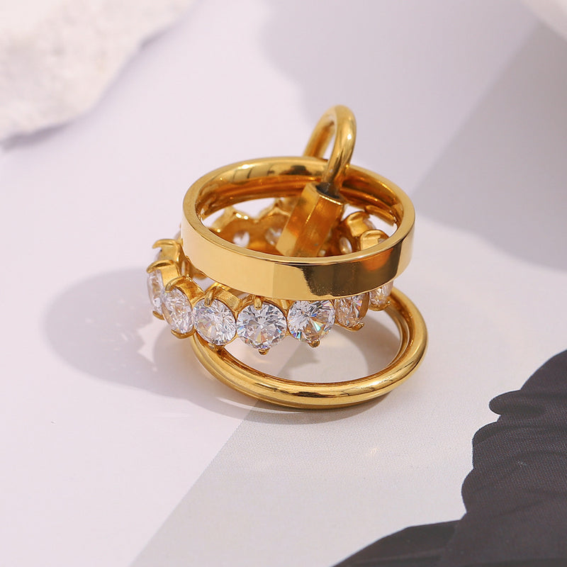 A stack of gold rings, including one with a row of clear round-cut stones, photographed on a light gray surface with soft shadows.