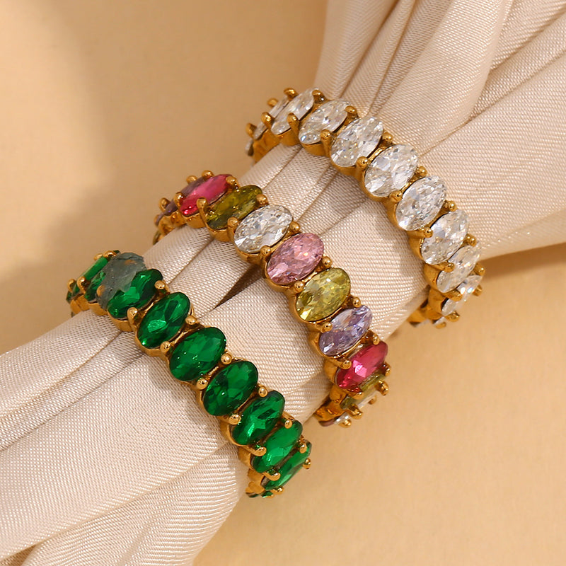 Three gold-plated bracelet cuffs with colorful gemstone accents, displayed on a cream-colored fabric wristband. The top bracelet features clear oval-cut stones, the middle features pink and yellow stones, and the bottom features green stones.