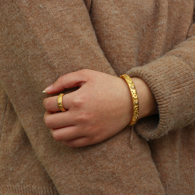 A close-up of a person's hand wearing a textured gold bangle bracelet and a matching gold ring, dressed in a cozy brown knit sweater.