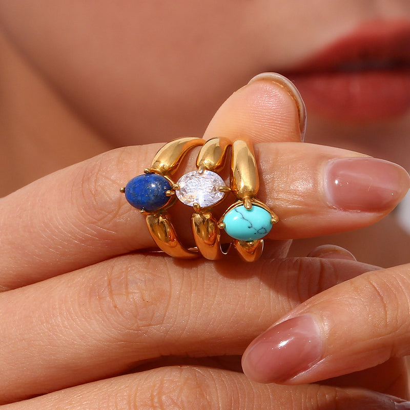 A close-up of  gold rings with three stacked bands, featuring a blue stone, a clear oval stone, and a turquoise stone, worn on a person's finger.