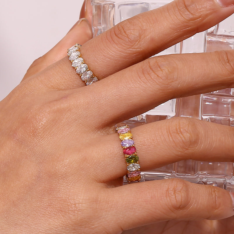 A close-up of a hand wearing two rings: a silver band with round clear stones on one finger and a colorful stone eternity band on the other. The colorful ring features segments of yellow, white, pink, green, and clear stones.