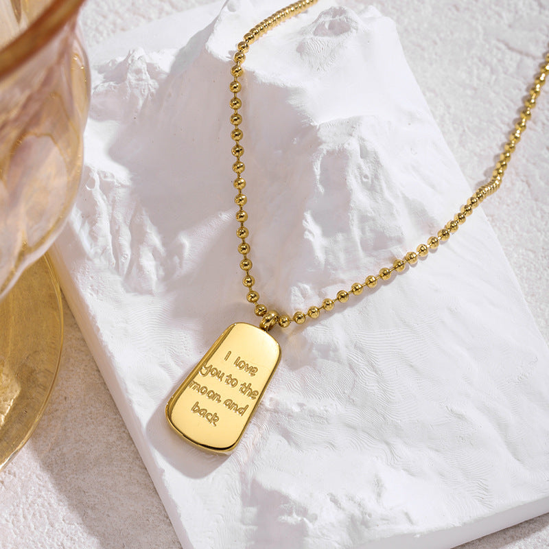A gold ball-chain necklace with a rectangular dog-tag-style pendant engraved with the phrase 'I love you to the moon and back,' styled on a textured white stone slab with a partial gold-toned glass visible at the left edge.