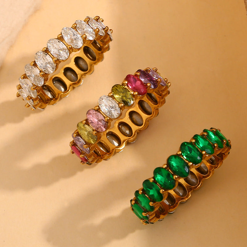 Three gold-plated stretch rings with oval-cut gemstones, displayed diagonally on a warm beige background. The top ring features clear stones, the middle ring has a mix of pink, green, and purple stones, and the bottom ring is lined with vibrant green stones.
