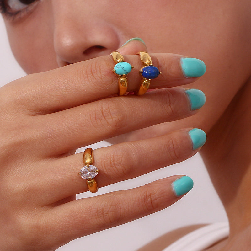 A close-up of a person's hand with turquoise nail polish wearing three gold rings: two with oval turquoise and blue cabochon stones, and one with a clear round-cut stone.