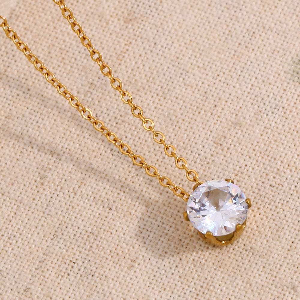 A gold chain necklace with a round clear faceted gemstone pendant in a gold setting, displayed on a textured beige fabric background.