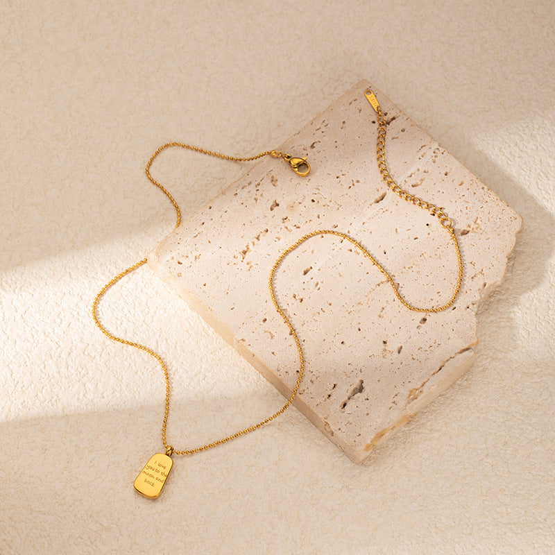 A delicate gold chain necklace with a small rectangular pendant engraved , styled on a textured beige stone slab against a warm beige background.