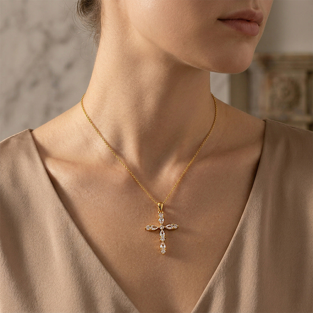 A close-up of a woman wearing a delicate gold cross pendant necklace adorned with small clear stones, paired with a beige satin top.
